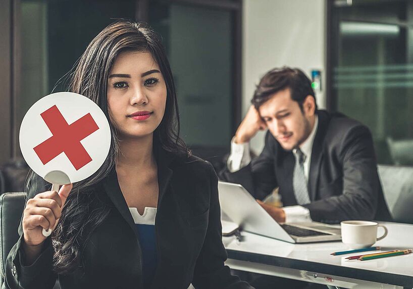 Frau hält Schild mit rotem Kreuz in die Höhe, Mann im Hintergrund schaut in einen Laptop