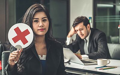 Frau hält Schild mit rotem Kreuz in die Höhe, Mann im Hintergrund schaut in einen Laptop